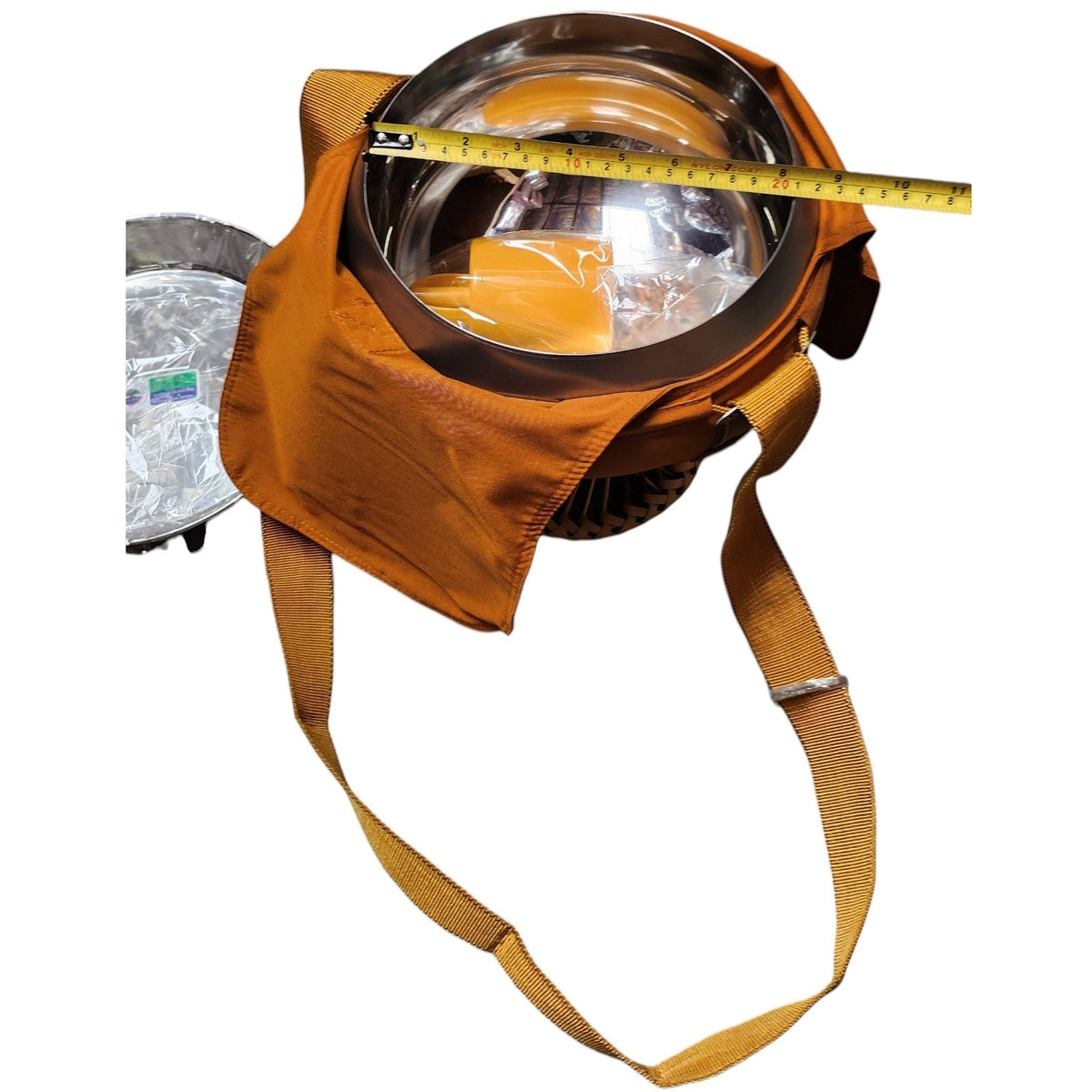 Alms bowl in orange cloth cover with a yellow tape measure stretched across the opening, steel plate at left
